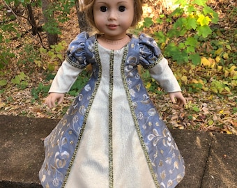 May include: A doll wearing a medieval-style dress. The dress features a light blue bodice with gold floral embroidery and gold trim. The skirt is cream-colored with a gold trim. The doll has blonde hair and is standing outdoors.