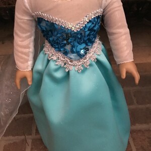 May include: A turquoise blue satin dress with a white mesh overlay and a silver and blue beaded bodice. The dress is designed for a doll.
