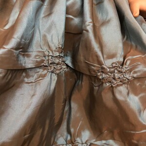 May include: A close-up of a sage green satin skirt with multiple layers and ruched detailing. The skirt has a gathered look and is made of a soft, flowing fabric.