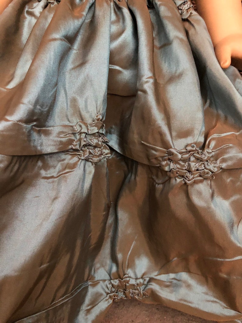 May include: A close-up of a sage green satin skirt with multiple layers and ruched detailing. The skirt has a gathered look and is made of a soft, flowing fabric.