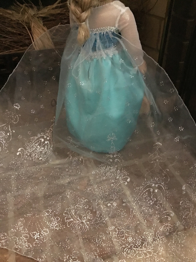 May include: A turquoise dress with a sheer white cape featuring a silver snowflake pattern. The dress has a blue bodice and a tulle skirt.