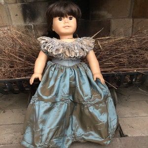 May include: A doll wearing a blue and brown dress with a large, ruffled collar. The doll has brown hair and brown eyes.