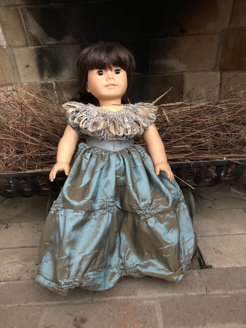 May include: A doll wearing a blue and brown dress with a large, ruffled collar. The doll has brown hair and brown eyes.