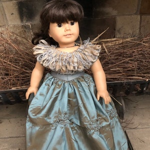 May include: A doll wearing a blue and brown dress with a silver and brown ruffled collar. The doll has brown hair and brown eyes.