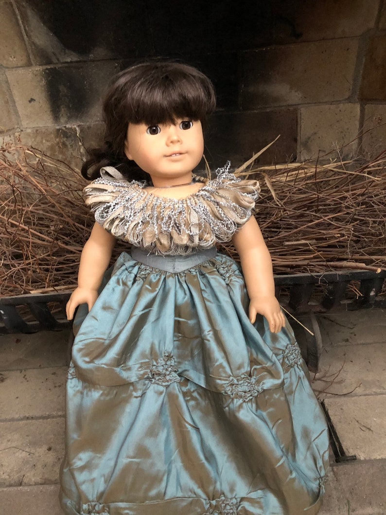 May include: A doll wearing a blue and brown dress with a silver and brown ruffled collar. The doll has brown hair and brown eyes.