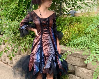 May include: A woman wearing a purple witch hat with a black veil and a long, iridescent dress with a black tulle overlay. She is wearing black lace-up boots.