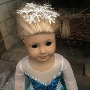 May include: A doll wearing a light blue dress with a white mesh overlay and a silver and blue trim. The doll has blonde hair and is wearing a white snowflake hair accessory.