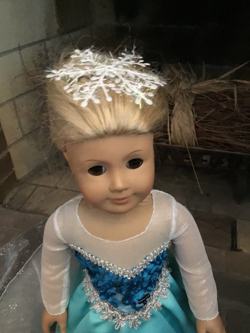 May include: A doll wearing a light blue dress with a white mesh overlay and a silver and blue trim. The doll has blonde hair and is wearing a white snowflake hair accessory.
