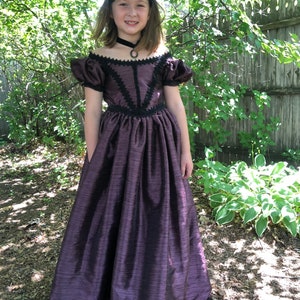 May include: A young girl wearing a purple dress with black lace trim and a black choker necklace. The dress has a full skirt and puffed sleeves. The girl is standing in a garden setting.