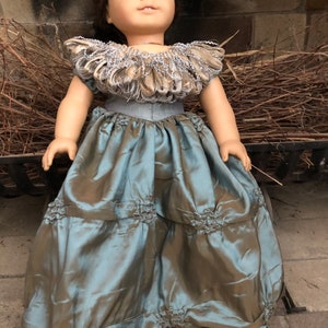 May include: A doll wearing a teal blue dress with a ruffled skirt and a brown and silver feathered collar.