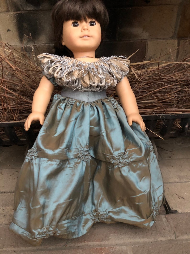 May include: A doll wearing a teal blue dress with a ruffled skirt and a brown and silver feathered collar.