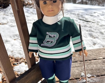 May include: A doll wearing a green and white hockey jersey with a large 'B' on the front. The doll is wearing blue pants and black skates. The doll is holding a hockey stick and is standing on a wooden deck with snow in the background.