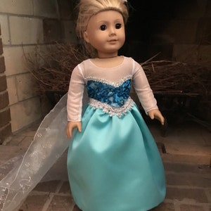 May include: A doll wearing a light blue and silver princess dress with a white sheer cape. The doll has blonde hair and brown eyes.