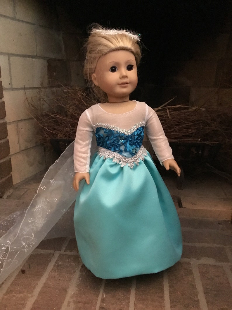 May include: A doll wearing a light blue and silver princess dress with a white sheer cape. The doll has blonde hair and brown eyes.