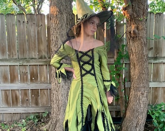 May include: A woman wearing a green witch costume with a black pointed hat and black boots. She is standing in front of a wooden fence and trees.