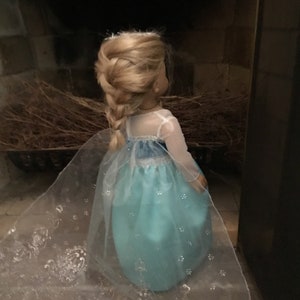 May include: A doll wearing a light blue dress with a white sheer overlay and a braid in her blonde hair. The dress has a snowflake pattern and the doll is standing in front of a dark background.