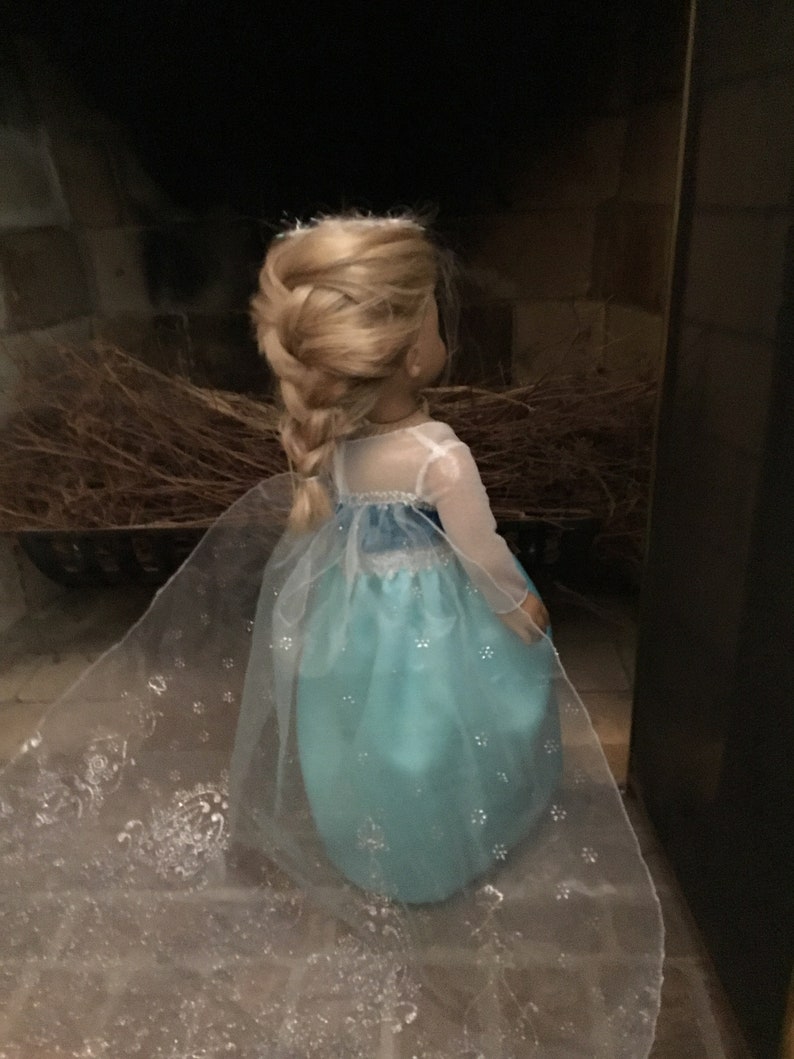 May include: A doll wearing a light blue dress with a white sheer overlay and a braid in her blonde hair. The dress has a snowflake pattern and the doll is standing in front of a dark background.