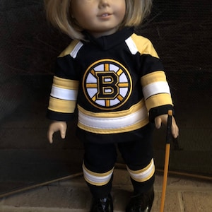 May include: A doll wearing a black and yellow hockey jersey with the Boston Bruins logo and a matching pair of pants. The doll is holding a wooden hockey stick.