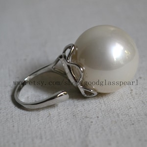 May include: A silver ring with a large, creamy-white pearl. The ring's band has a curved design, and the pearl is held in place by a decorative silver setting. The text "www.etsy.com/shop/goodglasspearl" is visible.