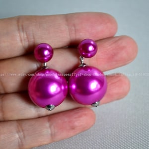 Puede incluir: Un par de pendientes de perlas de cristal rosa con postes de plata. Los pendientes son de doble capa con una perla más pequeña encima de una perla más grande.