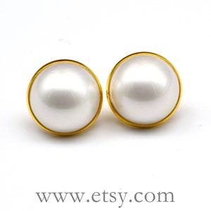 May include: A pair of large, round pearl earrings with a gold-colored border. The earrings have a bright, reflective surface and are set against a white background. The website address www.etsy.com is visible at the bottom.