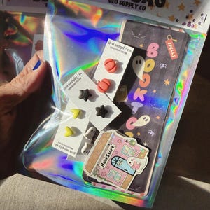 May include: A holographic Boo Bag from Geo Supply Co. containing several pairs of small stud earrings shaped like orange pumpkins, black stars, yellow hearts, and white ghosts, along with a bookmark labeled Spells and a bookstore sticker.