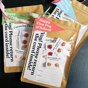 May include: Three brown paper pouches containing small stud earrings shaped like apples, goldfish, and pencils. Each pouch features a pennant tag reading Happy First Day of School and a card with the text Yay! Please return the card inside!