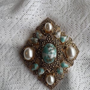 May include: A vintage gold-coloured brooch featuring an oval turquoise stone at its centre and four large faux pearls. The brooch has an ornate design with smaller turquoise and white stones.