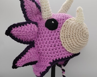 May include: Purple and white crocheted dinosaur hat with a black eye and a white horn. The hat has a long white and purple braided tie.