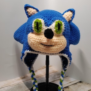 Könnte beinhalten: Blaue und weiße gehäkelte Sonic the Hedgehog Mütze mit grünen Augen und einer weißen und grünen geflochtenen Schnur.