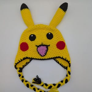 May include: A bright yellow crocheted hat featuring two long ears with black tips, two round red cheeks, black eyes with white highlights, and a pink tongue. The hat includes black trim along the bottom edge and two long braided yellow and black tassels.