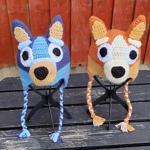 May include: Two crocheted dog hats, one blue and one orange, with black eyes and noses. The hats are on display stands.
