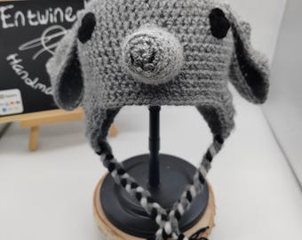 May include: Gray crocheted hat with a pig nose and black eyes. The hat has long gray, black, and white braided ties.