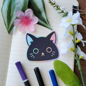 以下が含まれることがあります： 虹色の光沢のある黒猫のステッカー。白いノートに緑の葉と白い花が添えられています。