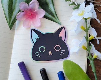 May include: A black holographic cat sticker with iridescent rainbow colors. The sticker is on a white notebook with green leaves and white flowers.