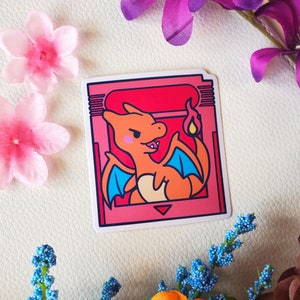 May include: A pink and blue cartoon sticker of a Charmander Pokemon character in a retro video game cartridge style.