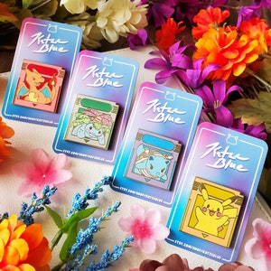 May include: Four enamel pins featuring Pokemon characters from the video game Pokemon Blue. Each pin is shaped like a Game Boy cartridge and features a different Pokemon: Charizard, Bulbasaur, Blastoise, and Pikachu. The pins are all gold-coloured with a white background and black outlines.