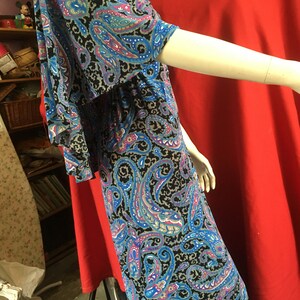 May include: A long, flowing dress with a paisley print in shades of blue, pink, and gray on a black background. The dress has a V-neckline and short sleeves.