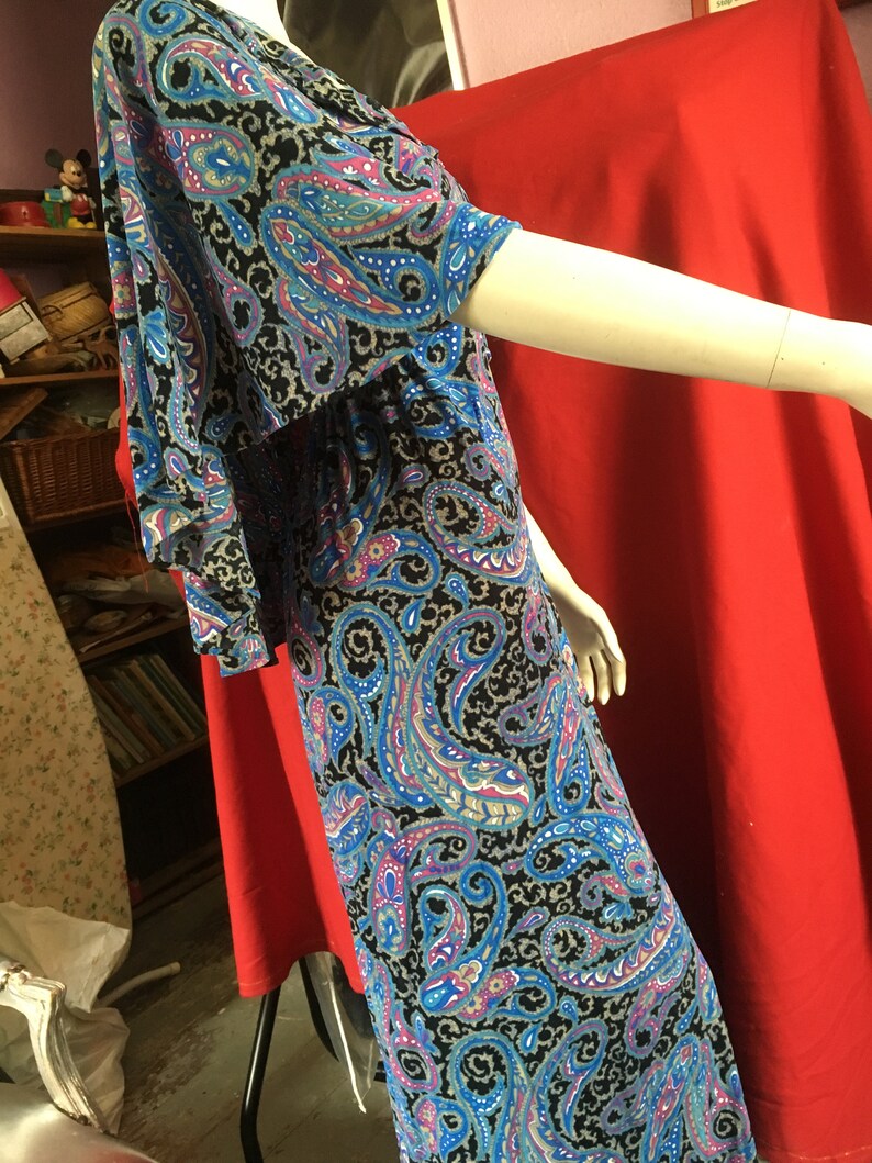 May include: A long, flowing dress with a paisley print in shades of blue, pink, and gray on a black background. The dress has a V-neckline and short sleeves.