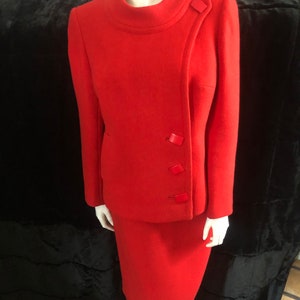 K&ouml;nnte beinhalten: Ein roter Wollanzug mit Rock und Jacke. Die Jacke hat einen hohen Kragen und einen diagonalen Verschluss mit vier roten Kn&ouml;pfen.