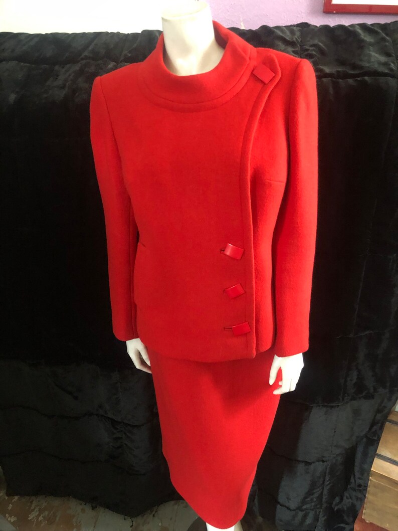 K&ouml;nnte beinhalten: Ein roter Wollanzug mit Rock und Jacke. Die Jacke hat einen hohen Kragen und einen diagonalen Verschluss mit vier roten Kn&ouml;pfen.