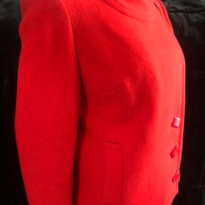 K&ouml;nnte beinhalten: Eine rote Wolljacke mit Stehkragen und drei roten Kn&ouml;pfen auf der Vorderseite. Die Jacke hat eine Tasche auf der linken Seite.