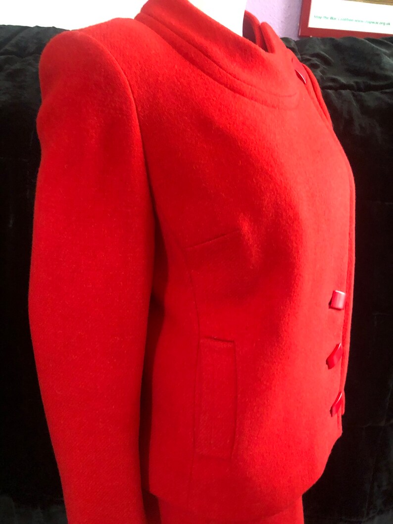 K&ouml;nnte beinhalten: Eine rote Wolljacke mit Stehkragen und drei roten Kn&ouml;pfen auf der Vorderseite. Die Jacke hat eine Tasche auf der linken Seite.