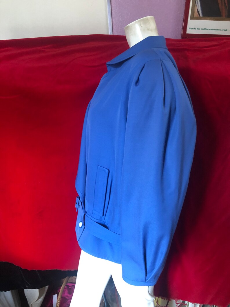May include: A blue jacket with a belted waist and a collar. The jacket has a slightly puffed sleeve and a single button closure.