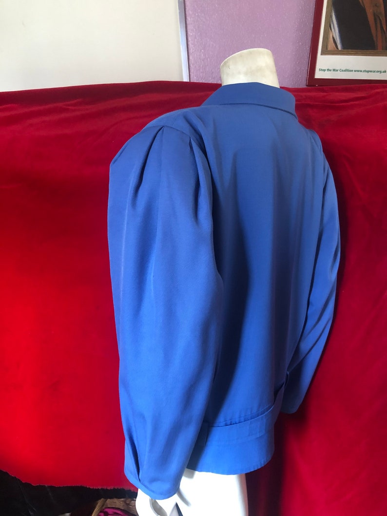 May include: A blue blazer with a long sleeve and a button-up collar. The blazer has a single button closure and a belt.