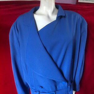 May include: A blue blazer with a long, asymmetrical collar, a belt, and two large buttons. The blazer is on a mannequin against a red background.