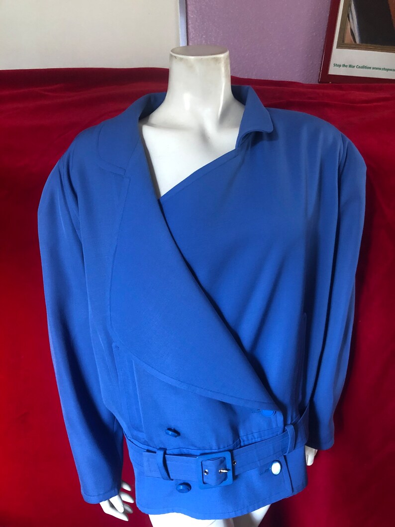 May include: A blue blazer with a long, asymmetrical collar, a belt, and two large buttons. The blazer is on a mannequin against a red background.