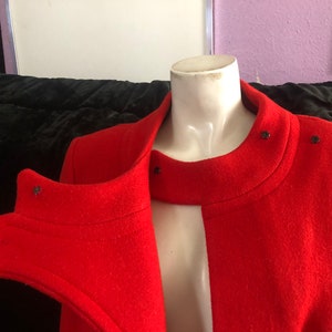 K&ouml;nnte beinhalten: Ein roter Wollmantel mit hohem Kragen und Knopfverschluss. Der Mantel hat ein einzigartiges Design mit einem geteilten Vorderteil und einem kontrastierenden schwarzen Besatz.