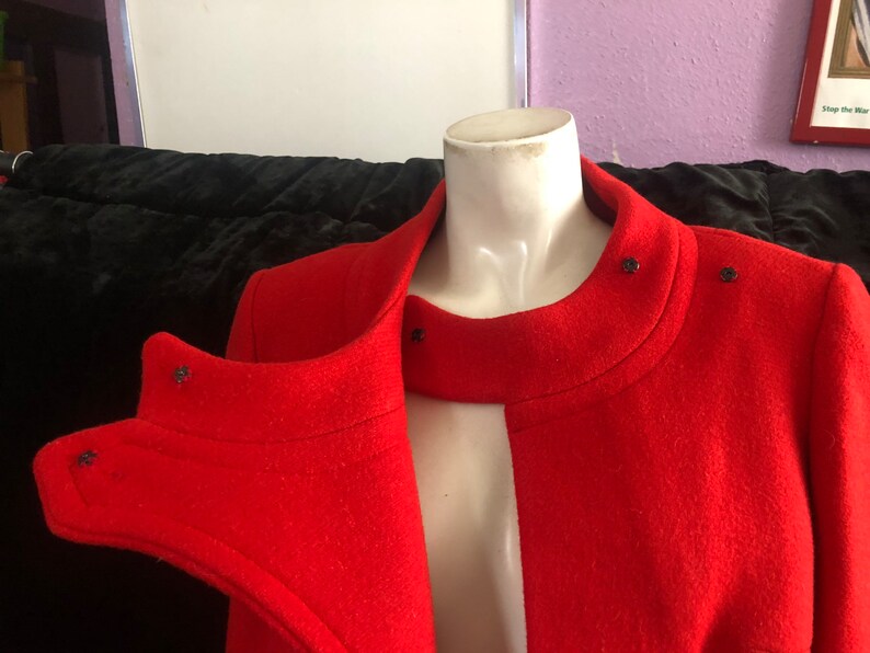 K&ouml;nnte beinhalten: Ein roter Wollmantel mit hohem Kragen und Knopfverschluss. Der Mantel hat ein einzigartiges Design mit einem geteilten Vorderteil und einem kontrastierenden schwarzen Besatz.