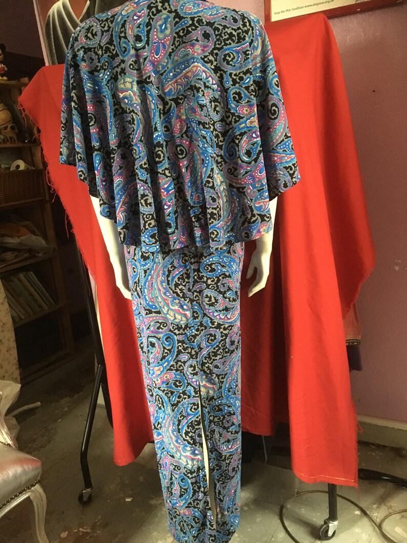 May include: A long, blue and black paisley print dress with a cape-like top. The dress has a slit up the back and is fitted through the bodice.